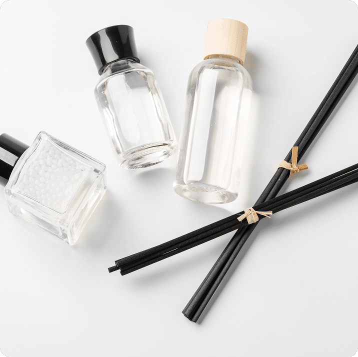 Three clear bottles with black and wooden caps are placed on a white surface alongside a bundle of black reed diffusers.